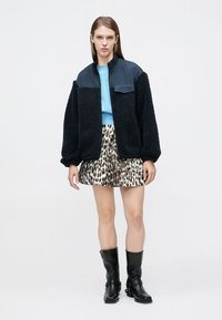 Μαύρο βελούδινο μπουφάν με σκούρο μπλε επάνω μέρος, συνδυασμένο με γαλάζιο τοπ και φούστα με σχέδιο λεοπάρ. Μαύρες μπότες μέχρι το γόνατο ολοκληρώνουν το σύνολο.