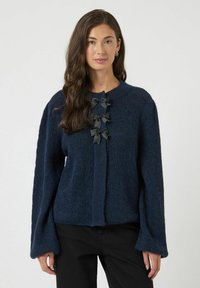 Maglione blu navy con tessuto strutturato, maniche lunghe e bottoni sul davanti, con tre fiocchi scuri come dettagli lungo la patta dei bottoni.