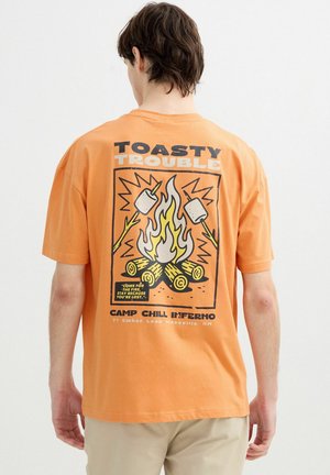 Hombre con camiseta naranja con gráfico de hoguera y malvaviscos y texto "Toasty Trouble" en la espalda, de pie frente a fondo blanco.
