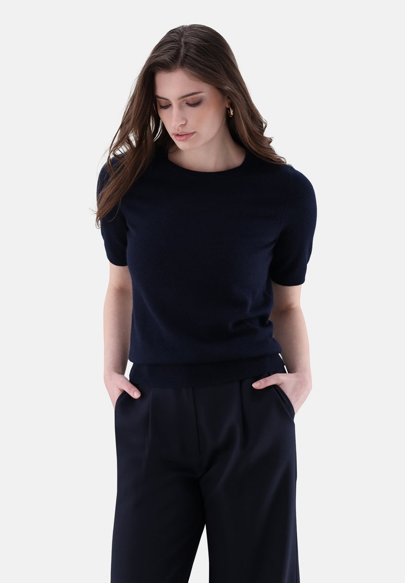 Navyblauer Kurzarmpullover aus weichem Material, Rundhalsausschnitt, figurbetonte Silhouette, kombiniert mit hochgeschnittenen dunklen Hosen, Hände in den Taschen.