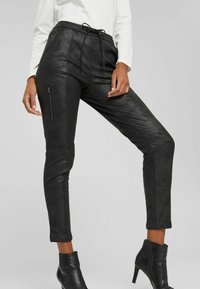 Black faux leather pants with a drawstring waist, fitted shape, zippered side pockets, and smooth texture. Finished with ankle-length hems.