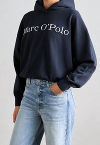 Sudadera con capucha azul marino con "Marc O'Polo" bordado en blanco, combinada con jeans de denim azul claro de corte clásico.