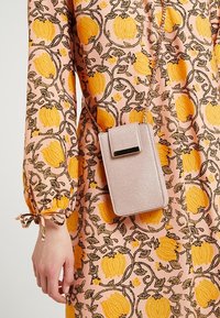 Jasnoróżowa torba na ramię o teksturowanym wykończeniu, z złotymi elementami ozdobnymi, klapkowym zapięciem i łańcuszkowym paskiem. Noszona z wzorzystą sukienką.