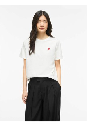 Jeune femme aux longs cheveux foncés portant un t-shirt blanc avec un petit cœur rouge et un pantalon noir plissé taille haute.