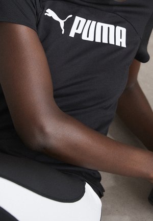 Nærbillede af en person iført en sort Puma-t-shirt og sort-hvide bukser, der viser deres arm og overkrop.