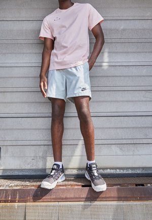 Hellrosa Baumwoll-T-Shirt mit Logo, graue Sportshorts und schwarze Laufschuhe mit grauen Akzenten; stehend auf einer strukturierten Oberfläche.