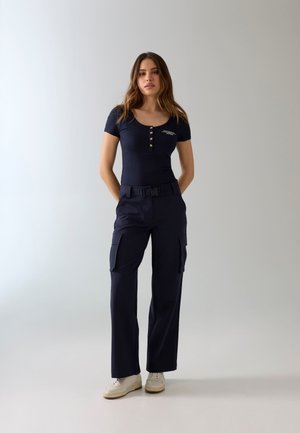 Jonge vrouw staande met handen achter de rug, draagt een navy blauw top met knopen, bijpassende cargobroek met riem en witte sneakers, neutrale achtergrond.