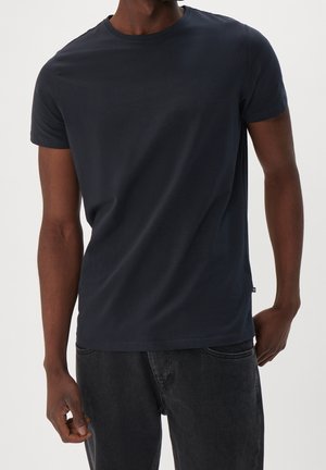 Mann trägt ein schlichtes dunkelblaues T-Shirt mit kurzen Ärmeln und schwarze Jeans, steht mit einer Hand in der Hosentasche vor einem neutralen Hintergrund.