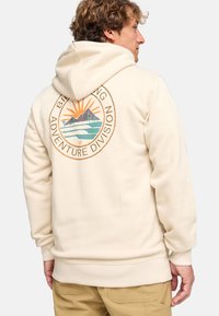 Sudadera beige hecha de tela suave con un logo en la parte posterior que presenta montañas y un sol, acentuado en naranja y verde.
