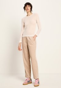 Frau steht mit der Hand in der Tasche ihrer beigen Hose, trägt ein figurbetontes, langärmliges Creme-Oberteil und rosafarbene Turnschuhe mit blauen Akzenten.