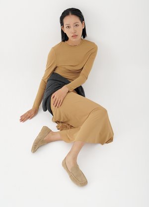 Mladá žena s krátkymi čiernymi vlasmi sedí na podlahe, oblečená v hnedohnedom dlhom rukáve, béžových mokasínach a tmavosivom svetri previazanom okolo pása.