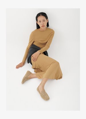 Fiatal nő rövid fekete hajjal, a padlón ülve, bézs hosszú ujjú ruhát, bézs mokaszint és sötétszürke pulóvert kötve a derekára visel.