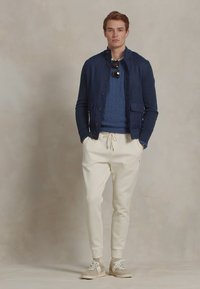 Navy gebreide cardigan over een blauwe gebreide trui, gecombineerd met witte joggerbroeken en beige sneakers. Accessoires zijn zwarte zonnebrillen.