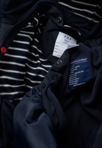 Den marineblå jakke har en indvendig foring med striber, en rød knap som accent og en plejeetiket, der detaljerer materialet og vejrbestandige egenskaber.