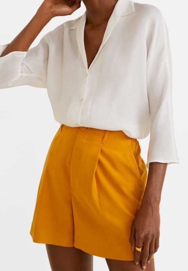 Personne portant un chemisier blanc à manches longues rentré dans un short jaune moutarde taille haute avec des plis et des passants de ceinture.