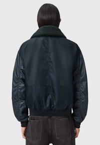 Henkilö, jolla on pitkät tummat hiukset ja tumma laivastonsininen bomber-takki, jossa on teksturoitu kaulus, seisoo selin yksinkertaista taustaa vasten.