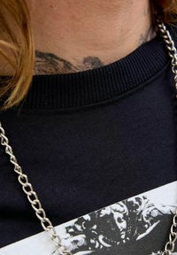 Sort sweatshirt med ribbet krave, der har et grafisk print. Synlig sølvkæde om halsen, baggrunden viser tatoveret hud.