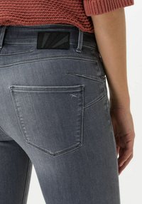 Jeans in denim grigio con un design aderente. Presentano due tasche posteriori e un'etichetta nera testurizzata in vita. Dettagli di cucitura discreti.