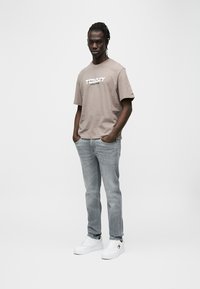 Καφέ βαμβακερό T-shirt με το λογότυπο "TOMMY JEANS" σε λευκό, συνδυασμένο με ξεθωριασμένο μπλε τζιν και λευκά αθλητικά παπούτσια. Ο μοντέλο στέκεται με τα χέρια στις τσέπες.