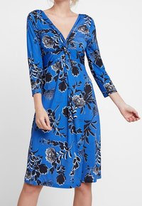 Donna che indossa un abito blu fino al ginocchio con motivi floreali in bianco e nero e maniche a tre quarti, caratterizzato da un dettaglio a nodo attorcigliato sul busto.