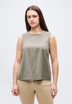 Γυναίκα με μακριά καστανά μαλλιά που φορά αμάνικο τοπ σε ελιά πράσινο και μπεζ παντελόνι, στέκεται μπροστά σε απλό φωτεινό φόντο.