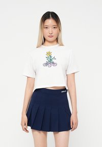 Tricou alb scurt cu un grafic de floare și panglică. Asortat cu o fustă clasică plisată bleumarin, cu un brâu elastic și detalii cu logo.