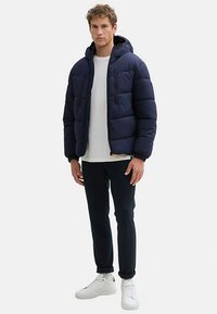 Granatowa pikowana kurtka z kapturem, kieszenie na zamek i ściągacze na mankietach. Nosi się ją na białą koszulkę, w połączeniu z ciemnymi spodniami i białymi trampkami.