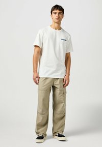 Camiseta blanca de algodón con logo azul de "Wrangler", combinada con pantalones cargo beige de corte holgado y zapatillas bajas en blanco y negro.