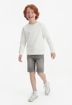 Rdečelasi otrok se smeji med hojo, oblečen v belo majico z dolgimi rokavi, sive denim kratke hlače in bele superge z visokimi konicami.