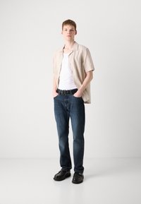 Light beige short-sleeve shirt over a white tank, paired with dark blue denim jeans and black shoes; relaxed fit and casual style.