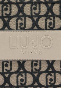 Béžová tkanina s opakujúcim sa čiernym vzorom loga a textúrovaným vzhľadom. Hladký, embosovaný kožený štítok nesie nápis "LIU·JO MILANO."