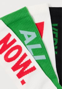 Três pares de meias: brancas com texto vermelho "AGORA", verdes com texto azul claro "TODAS" e pretas com texto verde "MUITO". Tecido tricotado, design canelado.