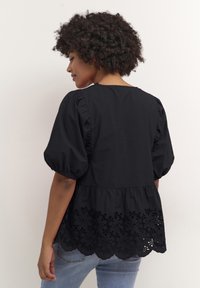 Črna majica s napihnjenimi rokavi, ki ima valovit rob in zapleteno izrezljan cvetlični vzorec na dnu. Mehka, teksturirana tkanina.