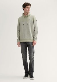 Felpa verde chiaro con corpo strutturato, cappuccio con coulisse e dettaglio del logo, abbinata a jeans scuri e sneaker nere.