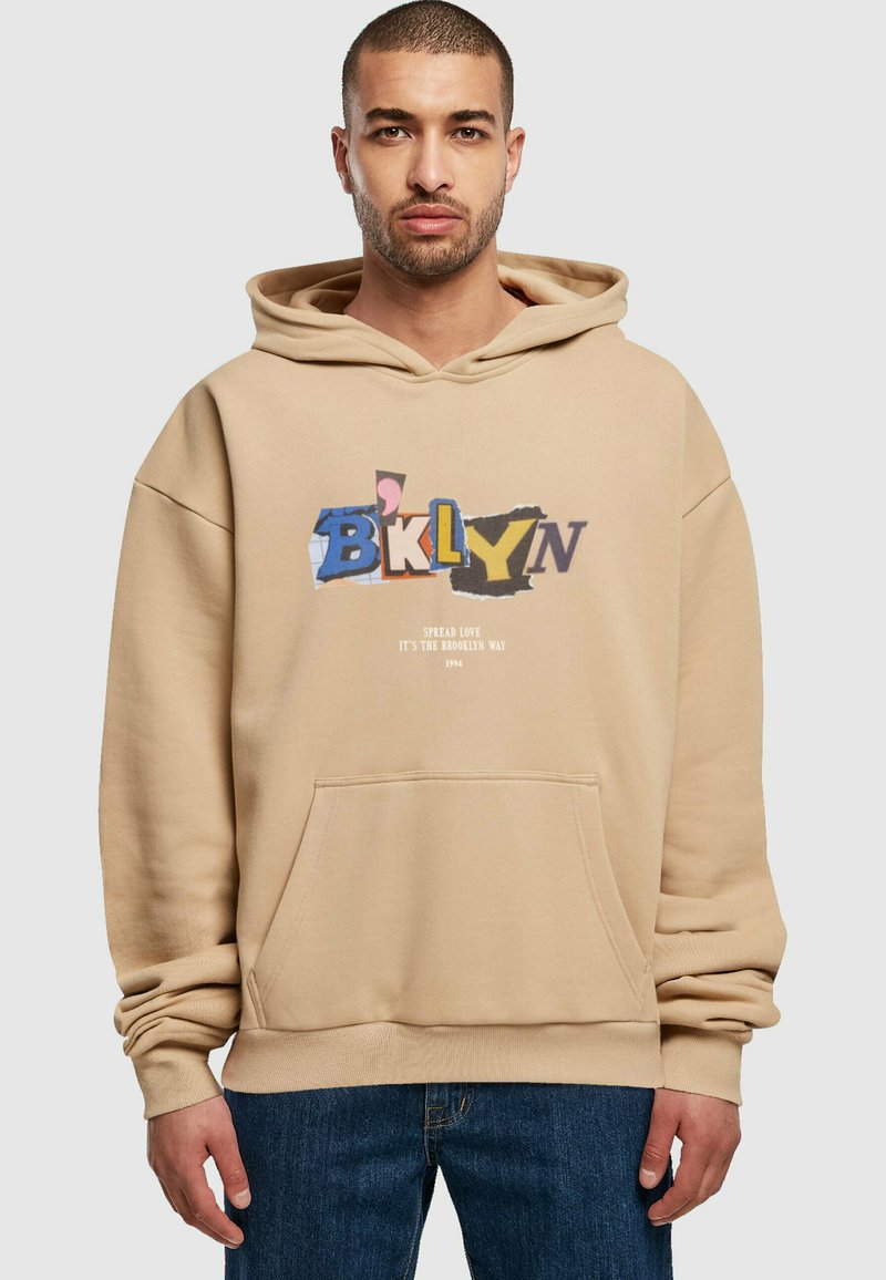 Sudadera beige con un bolsillo frontal. Presenta texto multicolor que dice "BKLYN" y "Spread Love It's the Brooklyn Way 1994". Textura suave.