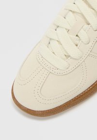 Zapatilla de ante color marfil con suela de goma texturizada, puntera redonda y cordones planos de algodón. Detalles de costuras realzan el diseño.