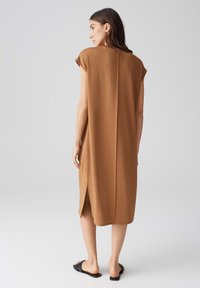 Braunes, knielanges Kleid mit kurzen Ärmeln, geradem Schnitt, Rückenschlitz und glatter Textur. Kombiniert mit schwarzen flachen Sandalen.