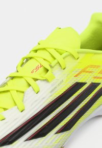 Εσωτερικό ποδοσφαιρικό παπούτσι σε φωτεινό κίτρινο με μαύρες και κόκκινες λεπτομέρειες, με υφή στην επιφάνεια και κορδόνια, σχεδιασμένο για βέλτιστη απόδοση.