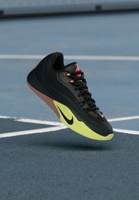 Μαύρο και νες κίτρινο αθλητικό παπούτσι Nike με πορτοκαλί λεπτομέρειες που αιωρείται πάνω από ένα μπλε αθλητικό γήπεδο με λευκές γραμμές.