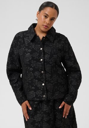 Femme aux cheveux foncés portant une veste noire à motif floral avec des boutons argentés et une jupe assortie, debout devant un fond gris uni.