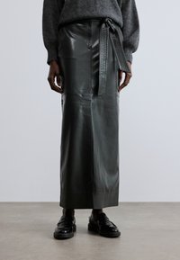 Roheline nahast midi seelik vöökohaga, sileda tekstuuri ja õmbledega, koos mustade loafer-kingadega neutraalsel taustal.