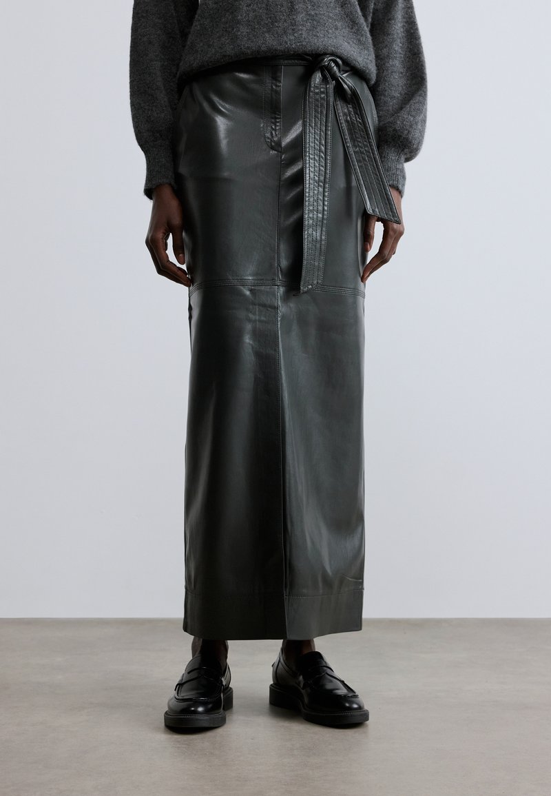 Roheline nahast midi seelik vöökohaga, sileda tekstuuri ja õmbledega, koos mustade loafer-kingadega neutraalsel taustal.