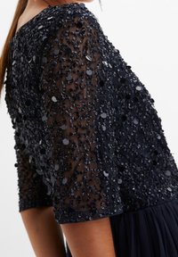 Marinblå klänning med genomskinliga, dekorerade ärmar som har paljetter och intrikata pärlbroderier. Flödande kjol under det dekorerade översta lagret.