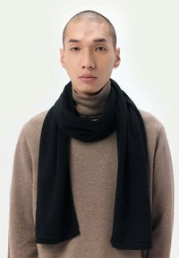 Svart scarf i mjukt material, draperad runt halsen, står i kontrast till en beige polotröja. Har en jämn textur och rena kanter.