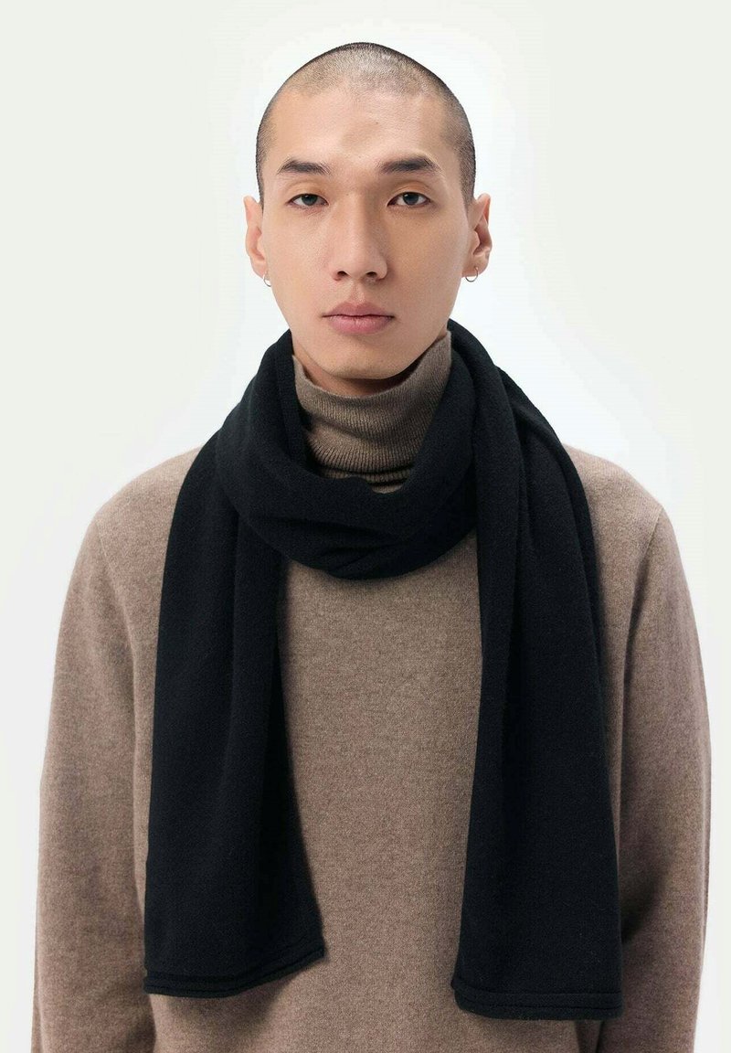 Svart scarf i mjukt material, draperad runt halsen, står i kontrast till en beige polotröja. Har en jämn textur och rena kanter.