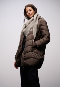 Brun pufferjakke med quiltet design, med et kontrasterende gråt mønstret tørklæde, båret med mørke jeans, stående mod en lys baggrund.