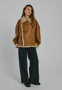 Vrouw in een bruine leren jas met schapenvacht, loszittende zwarte wijde jeans en zwarte schoenen, staand tegen een effen grijze achtergrond.