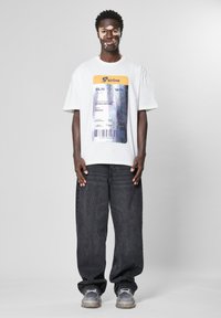 Hvit oversized t-skjorte med et fargerikt flybillettgrafikk. Kombinert med mørke baggy jeans og transparente joggesko. Enkel bakgrunn.