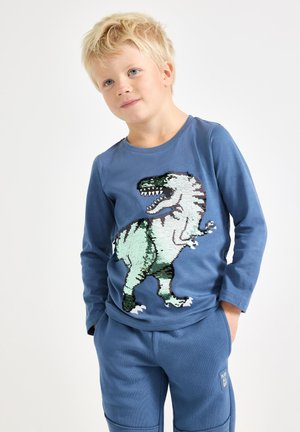 Plava majica dugih rukava s dizajnom dinosaurusa u zelenoj i crnoj boji sa šljokicama. Mekani materijal uz odgovarajuće plave hlače. Ležerna dječja odjeća.