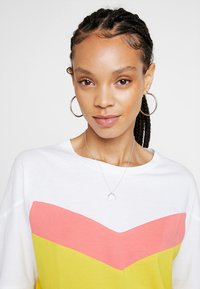 Camiseta blanca con un diseño a bloques de color que presenta patrones de chevron en rosa y amarillo, combinada con aretes de aro y un collar delicado con un colgante.